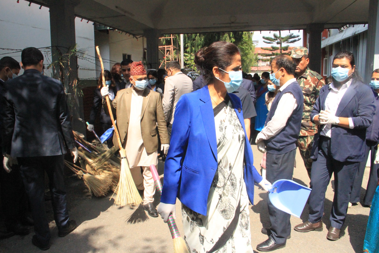 मास्क र पन्जा लगाएका मानिसहरूले बाहिर सामूहिक सफाई प्रयासमा भाग लिन्छन्, ढाकिएको पैदल मार्गमुनि झाडु, डस्टप्यान र सफाई उपकरणहरू बोकेर।